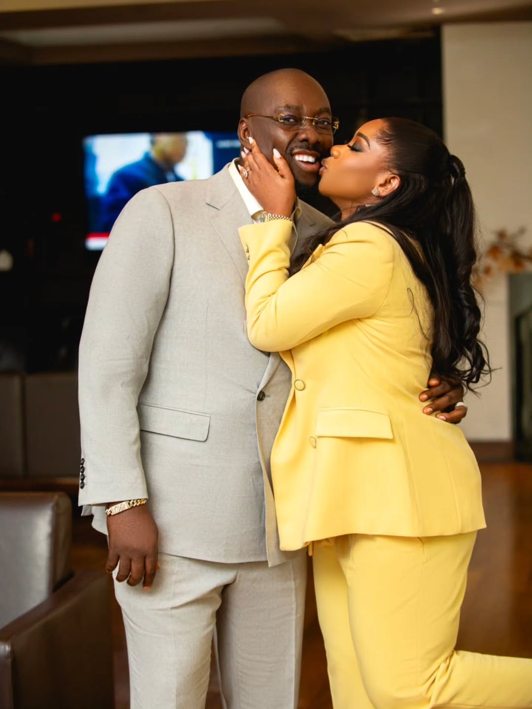 Ebele Iyiegbu in a butter yellow pantsuit affectionately kissing her husband Obi Cubana, who is wearing a stone grey suit, on the cheek.