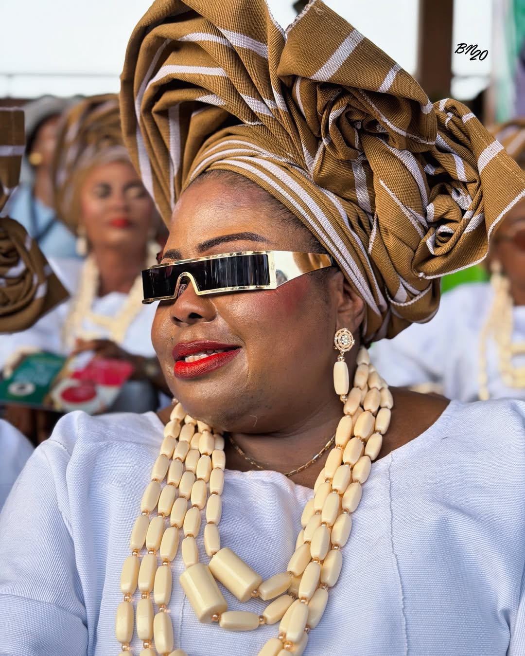 An attendee at the Ojude Oba festival wearing structured gold-rimmed rectangular sunglasses, a gold-striped Aso Oke gele, and heavy ivory beads.