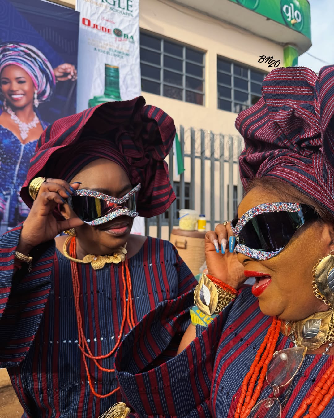 Two women in wine and blue striped Aso Oke at Ojude Oba 2026 wearing wide-lens shield sunglasses bordered with dense multi-coloured glitter.