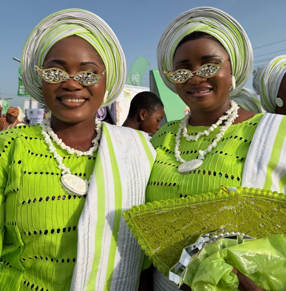 Two Ijebu-Ode women smiling at the Ojude Oba Festival wearing matching lime green buba, white and green gele, and stone-embellished sunglasses.