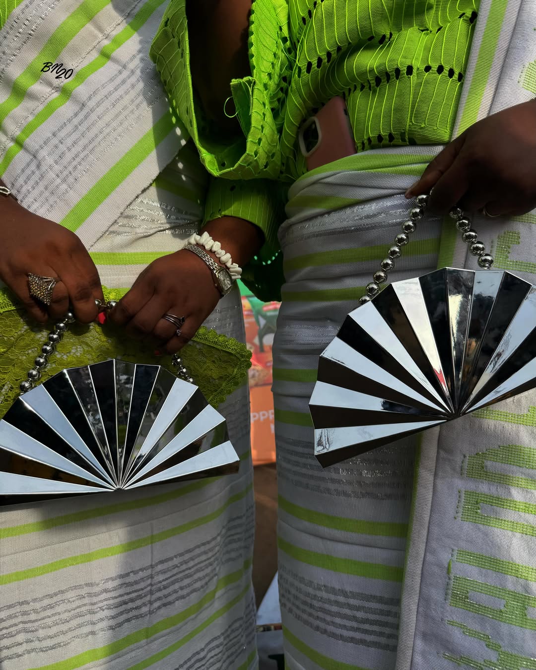 Two women holding identical structured silver metallic fan-shaped purses while wearing lime green and white buba and iro.