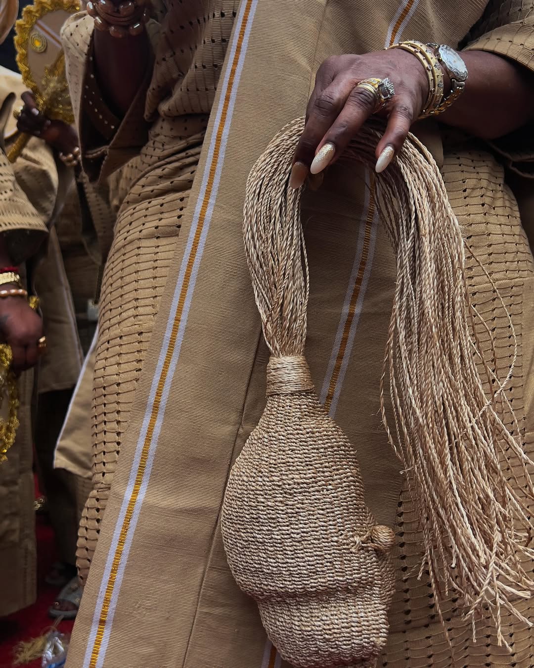 Close-up of a woman's hands with white manicured nails holding a custom woven beige horse-tail style purse over a matching aso-oke outfit.