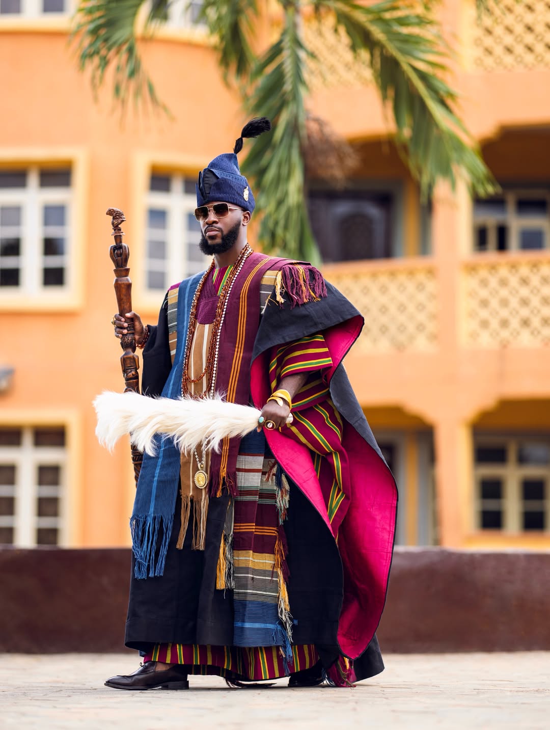 Akin Faminu standing outdoors at Ojude Oba 2026 wearing a multi-toned wine, gold, and green striped Aso-Oke robe with a magenta-lined black cloak, holding a traditional staff and white horse-tail iruekere.