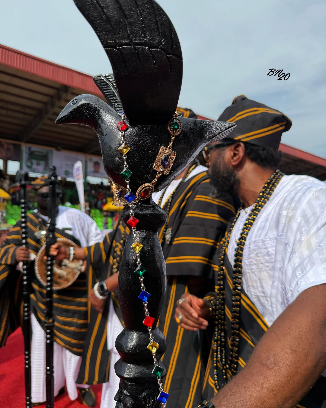 A carved black traditional staff topped with a bird figure, wrapped in multi-coloured rhinestones, held by a man in an agbada.