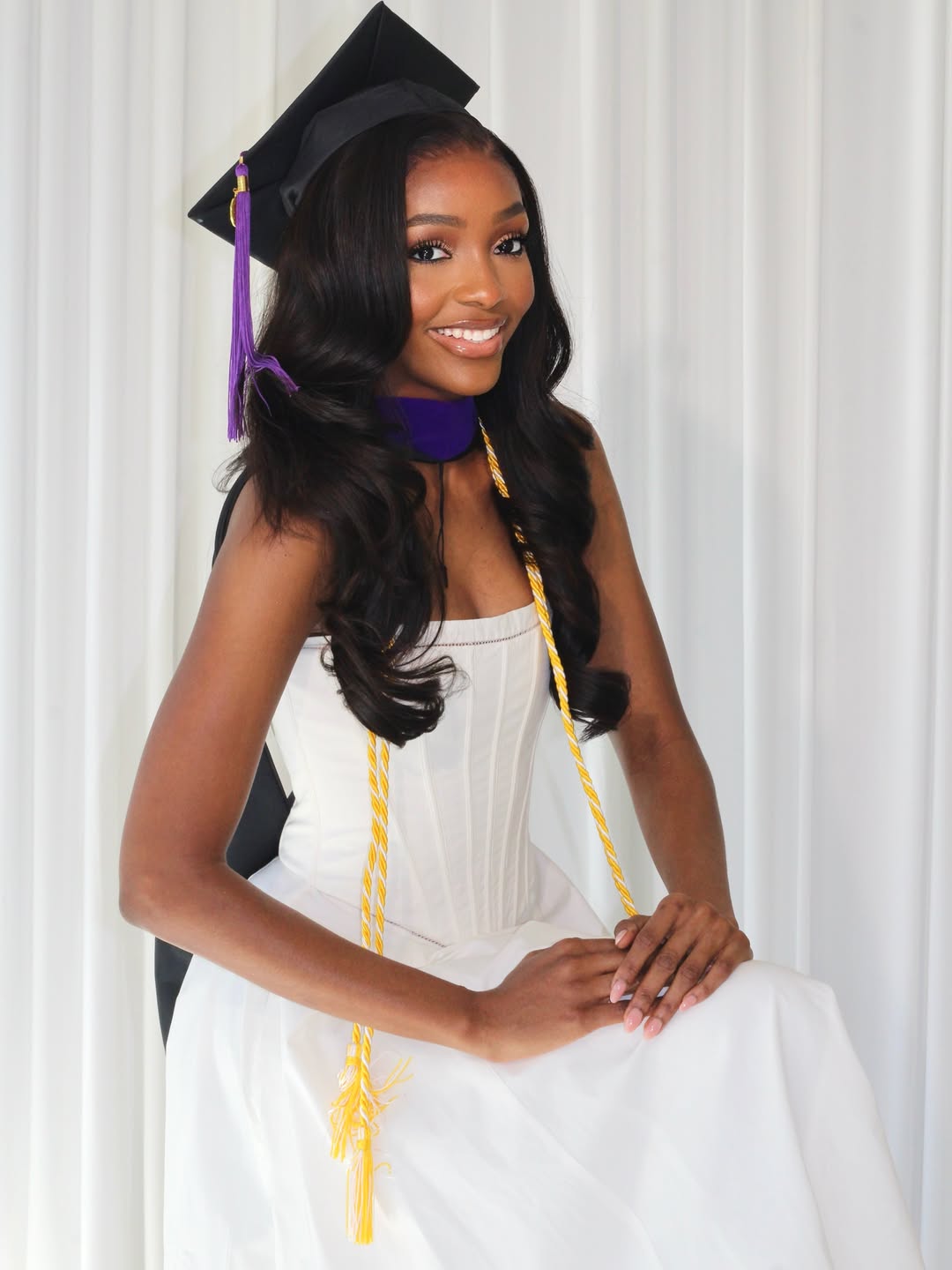 The 40th Miss Nigeria Chioma Amadi seated in a white corset dress with yellow graduation honor cords and a purple academic hood.