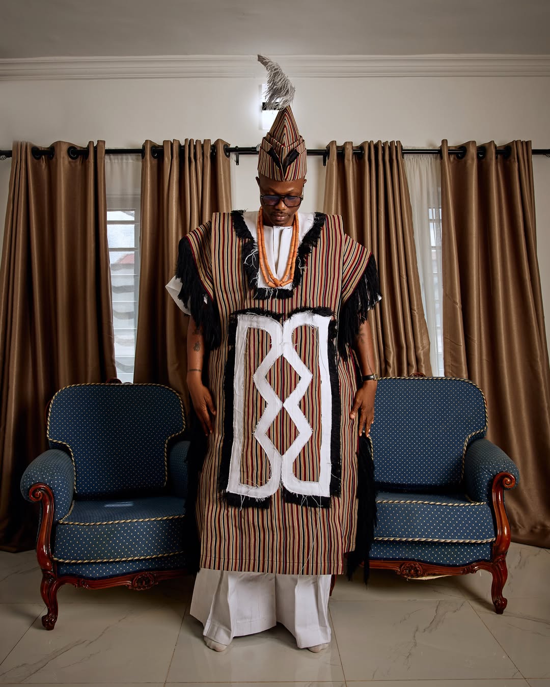 Vector posing indoors in a structured, sleeveless striped beige and brown traditional robe featuring a bold geometric monochrome center panel, black shoulder fringe, and a plumed fila cap.