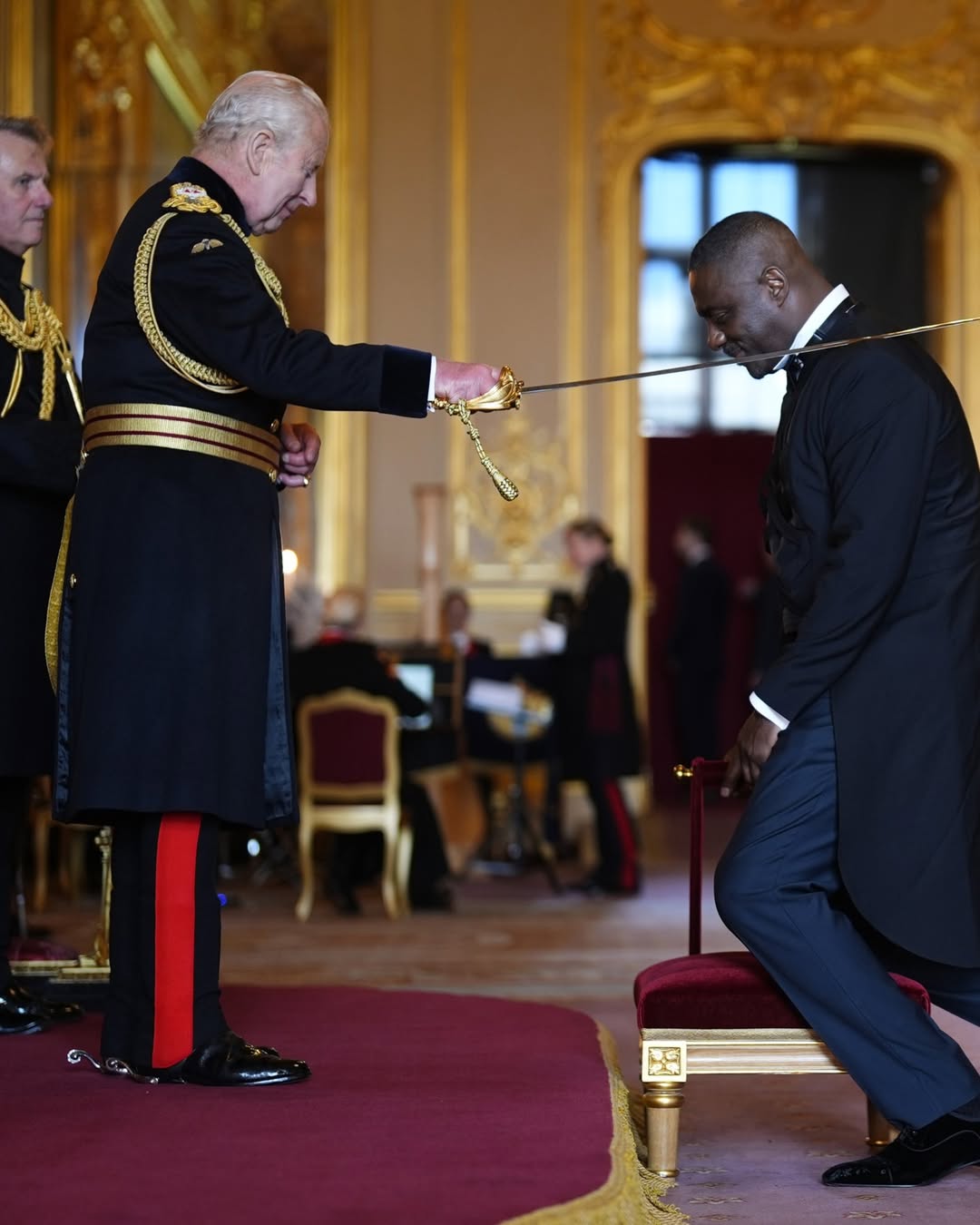 Idris Elba kneeling on a red velvet stool as King Charles III touches his shoulder with a ceremonial sword during his knighthood investiture ceremony inside Windsor Castle.