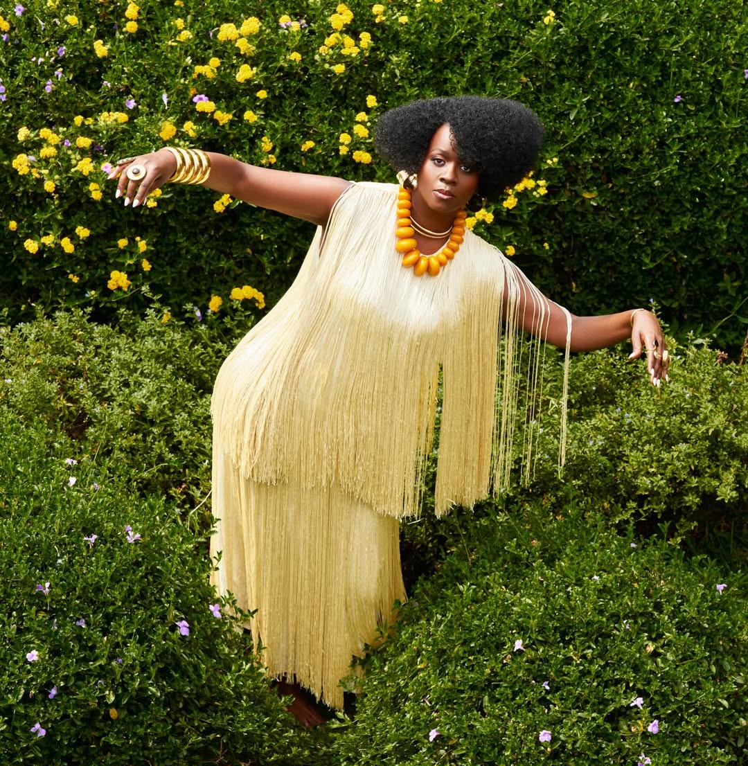 Singer Somi poses with outstretched arms in a cream fringe dress and large orange beads amidst green bushes with yellow flowers.