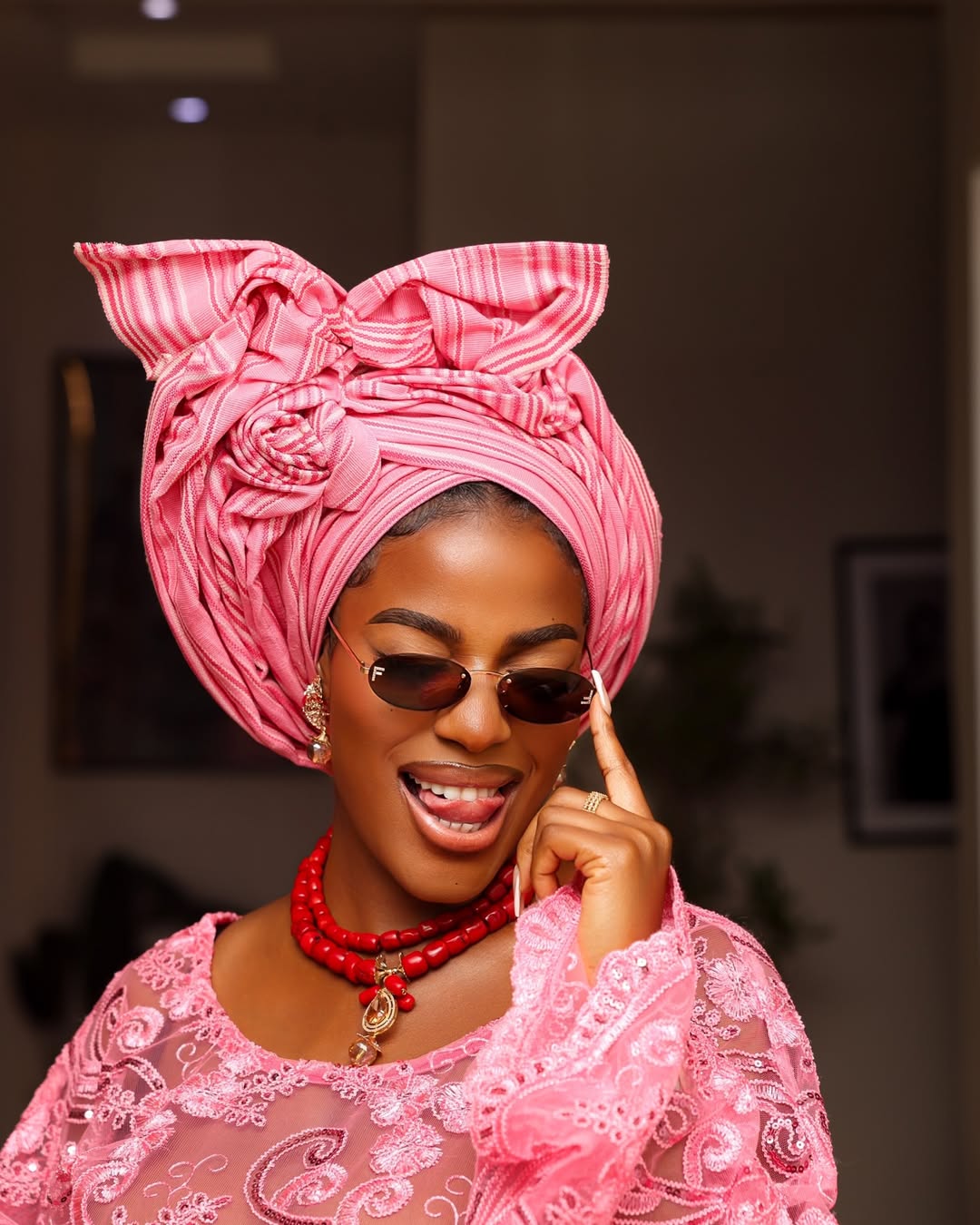 Close-up of culinary icon Hilda Baci highlighting her meticulously tied pink aso-oke gele with sculptural architectural folds, layered traditional red coral beads, and retro sunglasses.