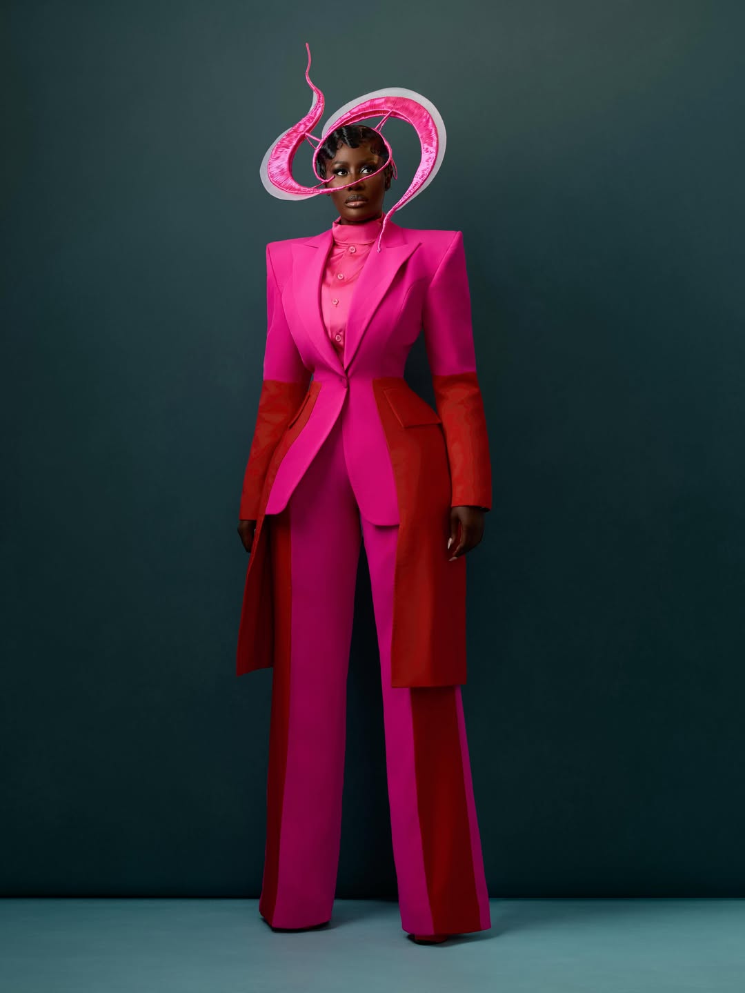 Nana Akua Addo posing in a structured hot pink and deep red tailored blazer and trouser suit by Mimi Yina of Medlin Boss, accessorised with an abstract organic pink and white sculptural headpiece.