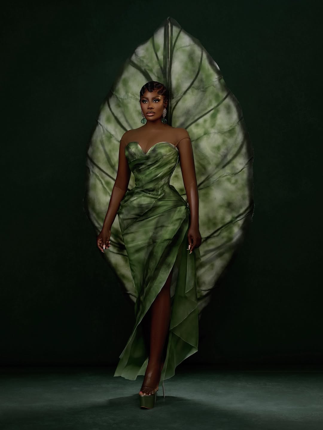 Nana Akua Addo in a strapless forest green corseted gown with a high leg split by Mohammed Abbas Ossu, posing against a matching large illuminated green leaf backdrop.