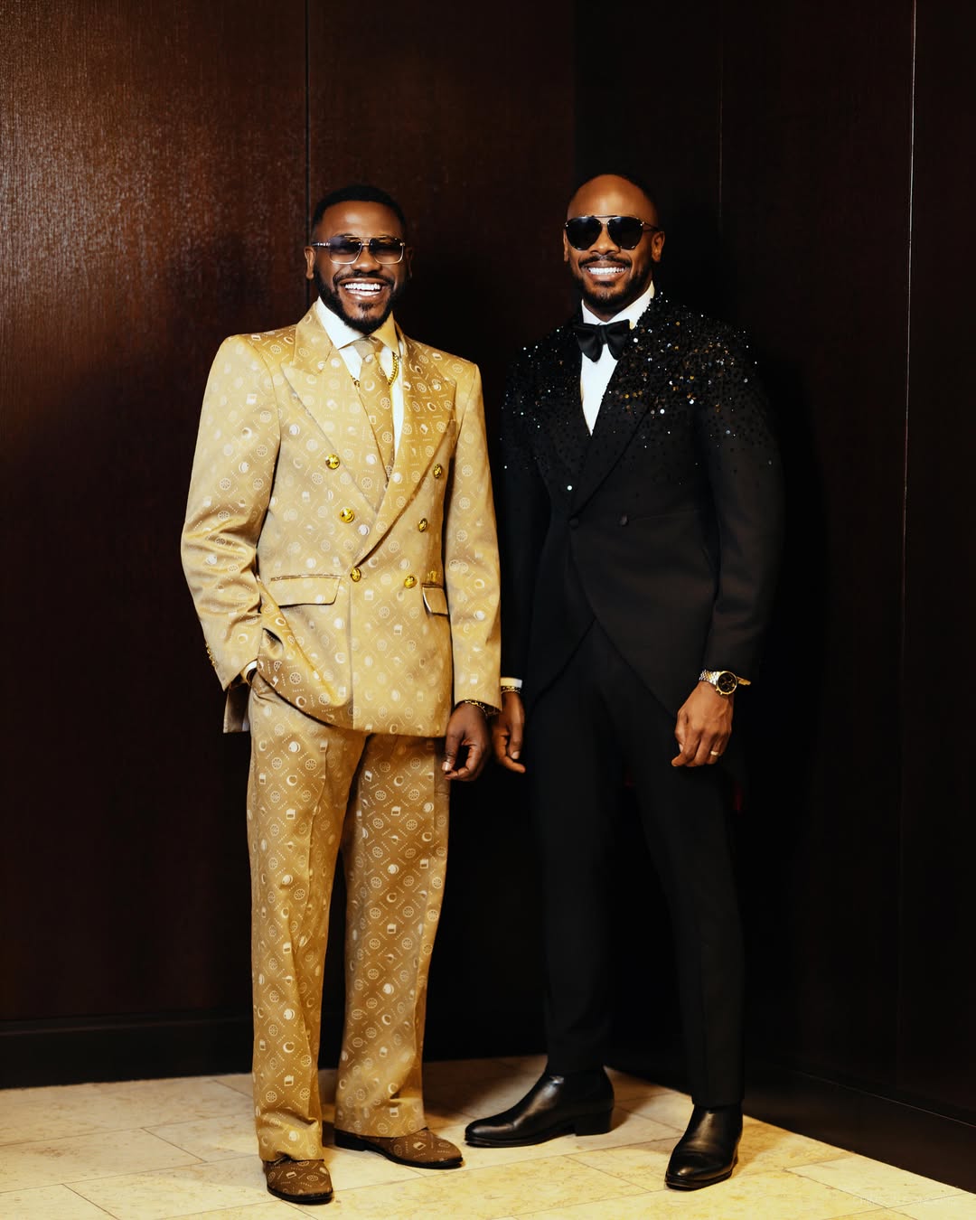 Actors Deyemi Okanlawon in a textured gold double-breasted suit and Daniel Etim Effiong in an embellished black tailcoat tuxedo posing together against a dark wooden backdrop.