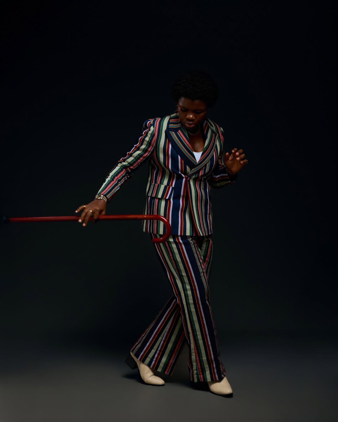 Professional dancer Endurance Grand posing against a dark backdrop in a multi-coloured vertical striped blazer and matching wide-leg trousers, holding a reddish-brown walking cane.