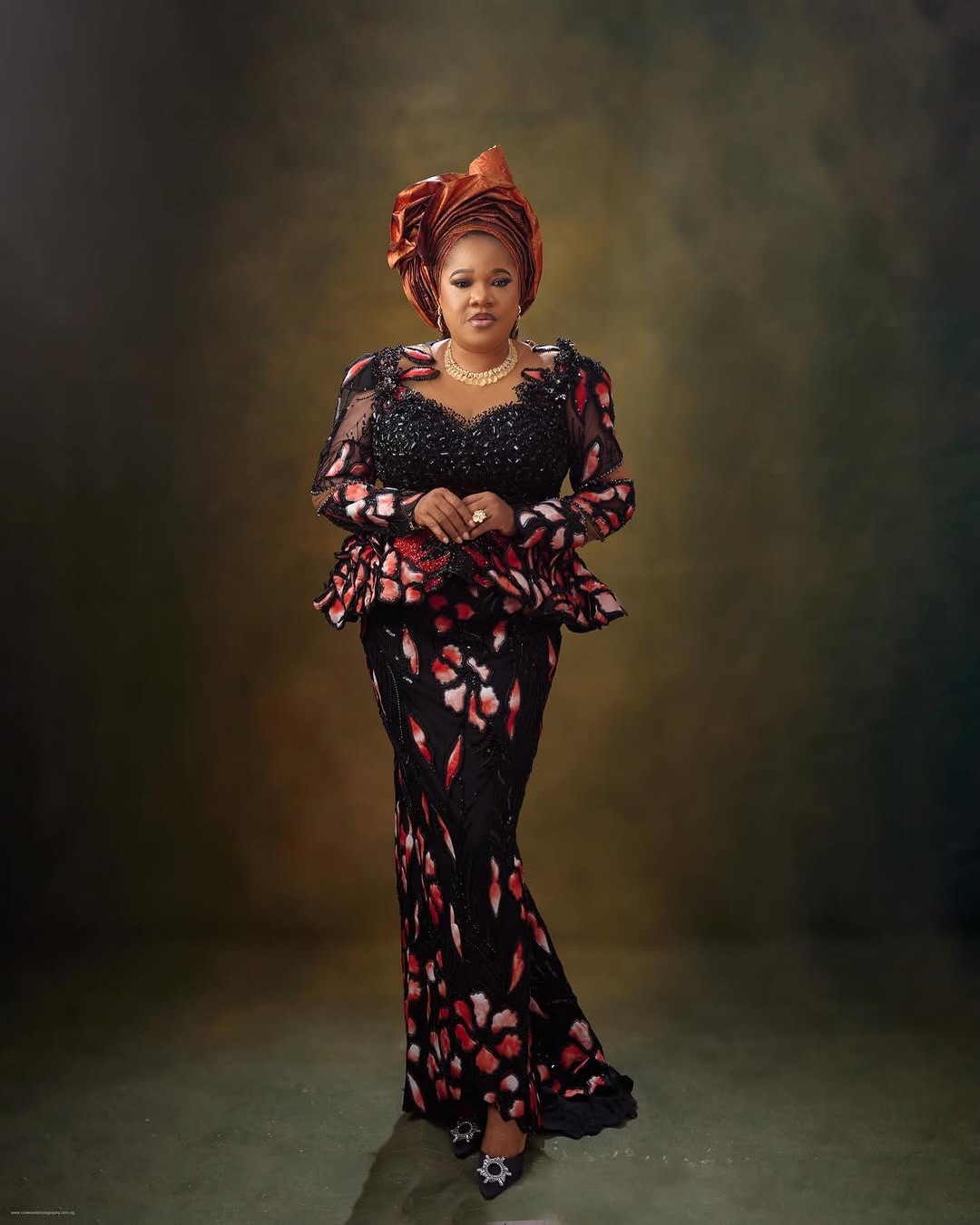 Toyin Abraham Ajeyemi walking forward looking down, wearing a copper architectural headtie and crystal-buckled heels.