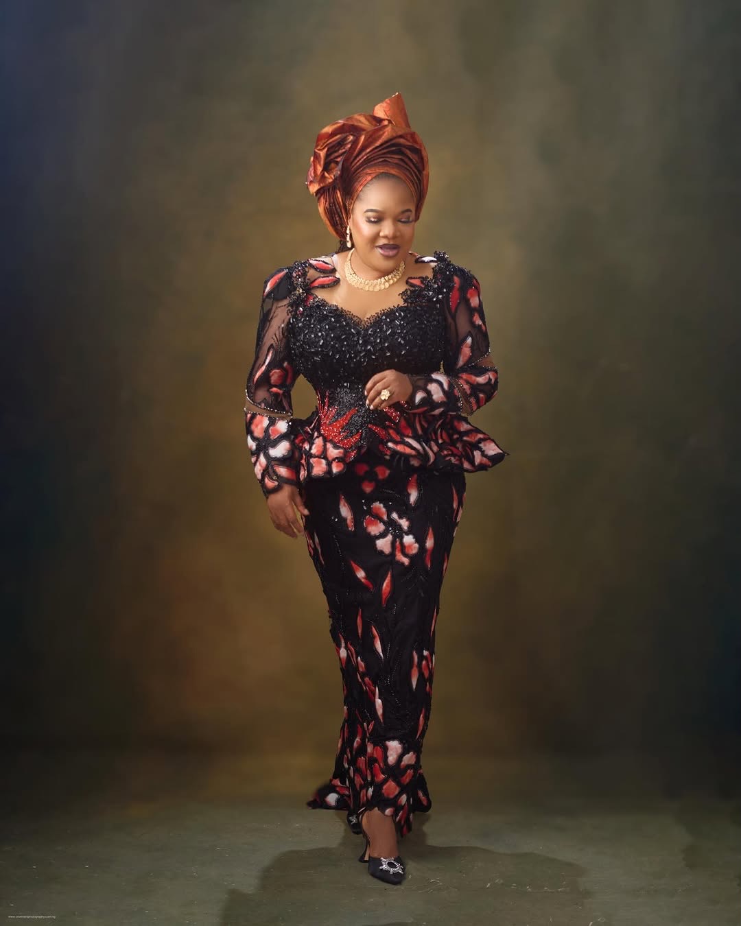 Full-length view of Toyin Abraham Ajeyemi standing with hands clasped in a black and orange floral lace skirt and blouse.