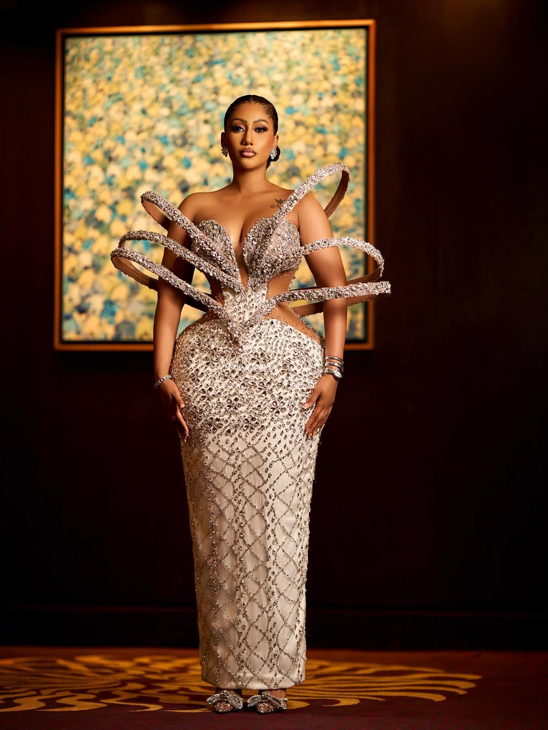 Mona against a dark wall in a strapless cream evening gown completely covered in multi-sized crystals, framed by an architectural cage-like loop structure.