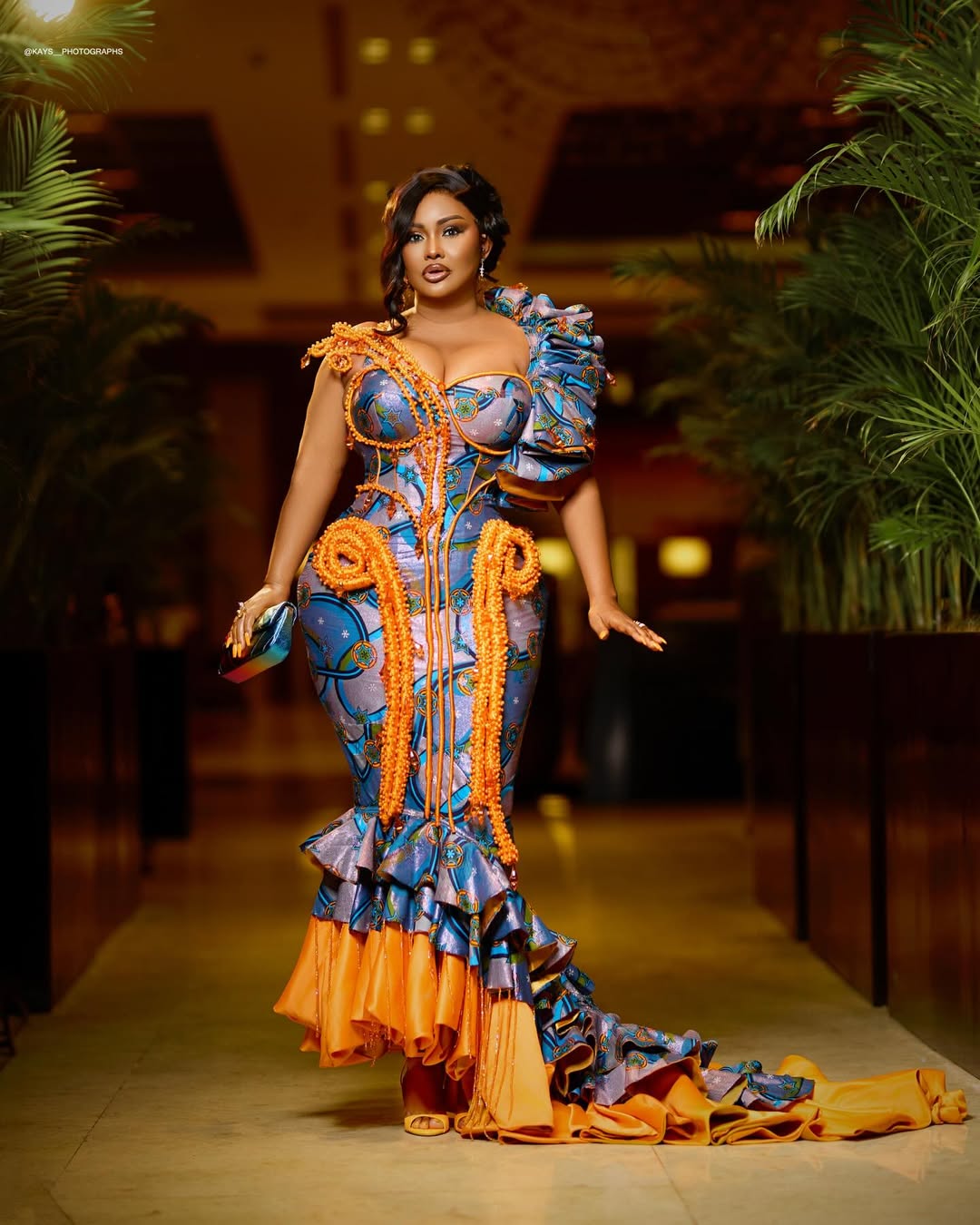 Ghanaian actress Nana Ama McBrown posing in a custom structured fishtail gown made from patterned blue ankara fabric, detailed with orange beadwork and tiered ruffles at the hem.