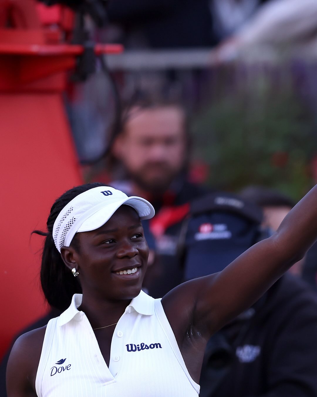 Canadian teenage tennis player Victoria Mboko smiling and waving to the crowd at the Queen's Club Championships while wearing a white Wilson visor and top.