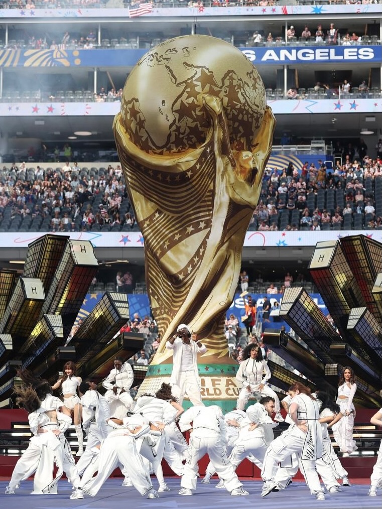 Rema, Lisa, Anitta, and a large group of dancers perform on the stadium field in front of a giant inflatable gold FIFA World Cup trophy.