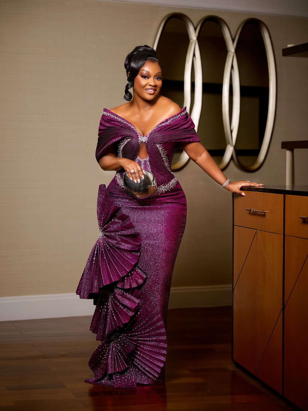 Jackie Appiah smiling and leaning against a wooden cabinet, showcasing the off-the-shoulder portrait neckline and rhinestone trim of her purple gown.