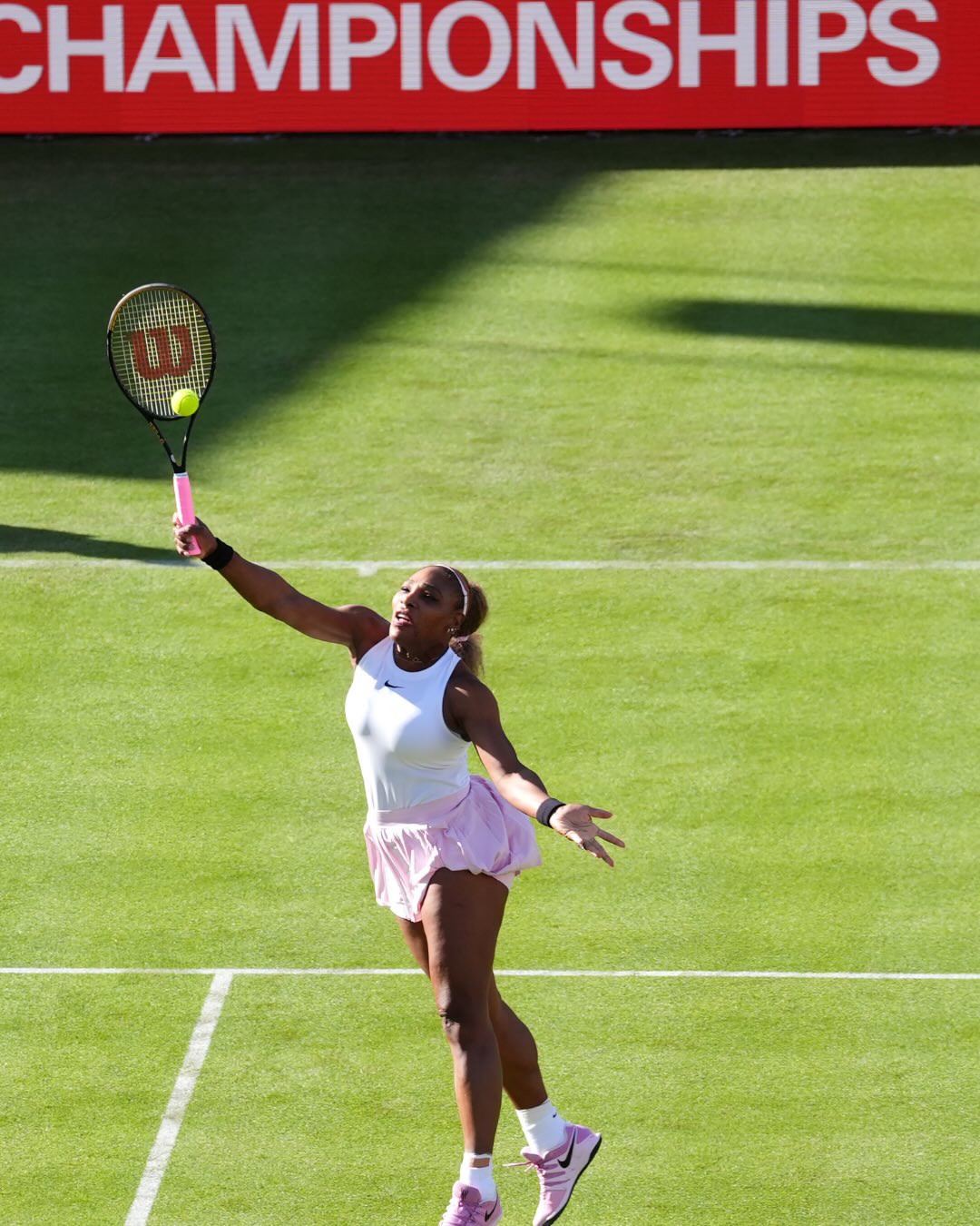 Serena Williams leaping on the green grass court to hit an overhead smash with her Wilson racket beneath the Queen's Club signage.