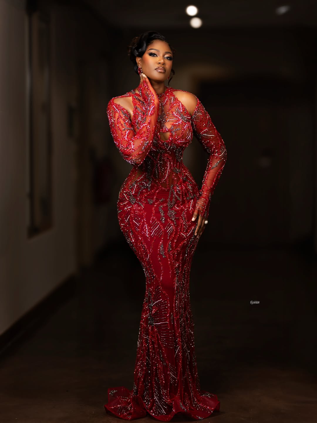 Actress Yvonne Nelson standing in a dimly lit hallway wearing a floor-length crimson red column gown on sheer tulle, intricately detailed with silver and metallic beadwork patterns.