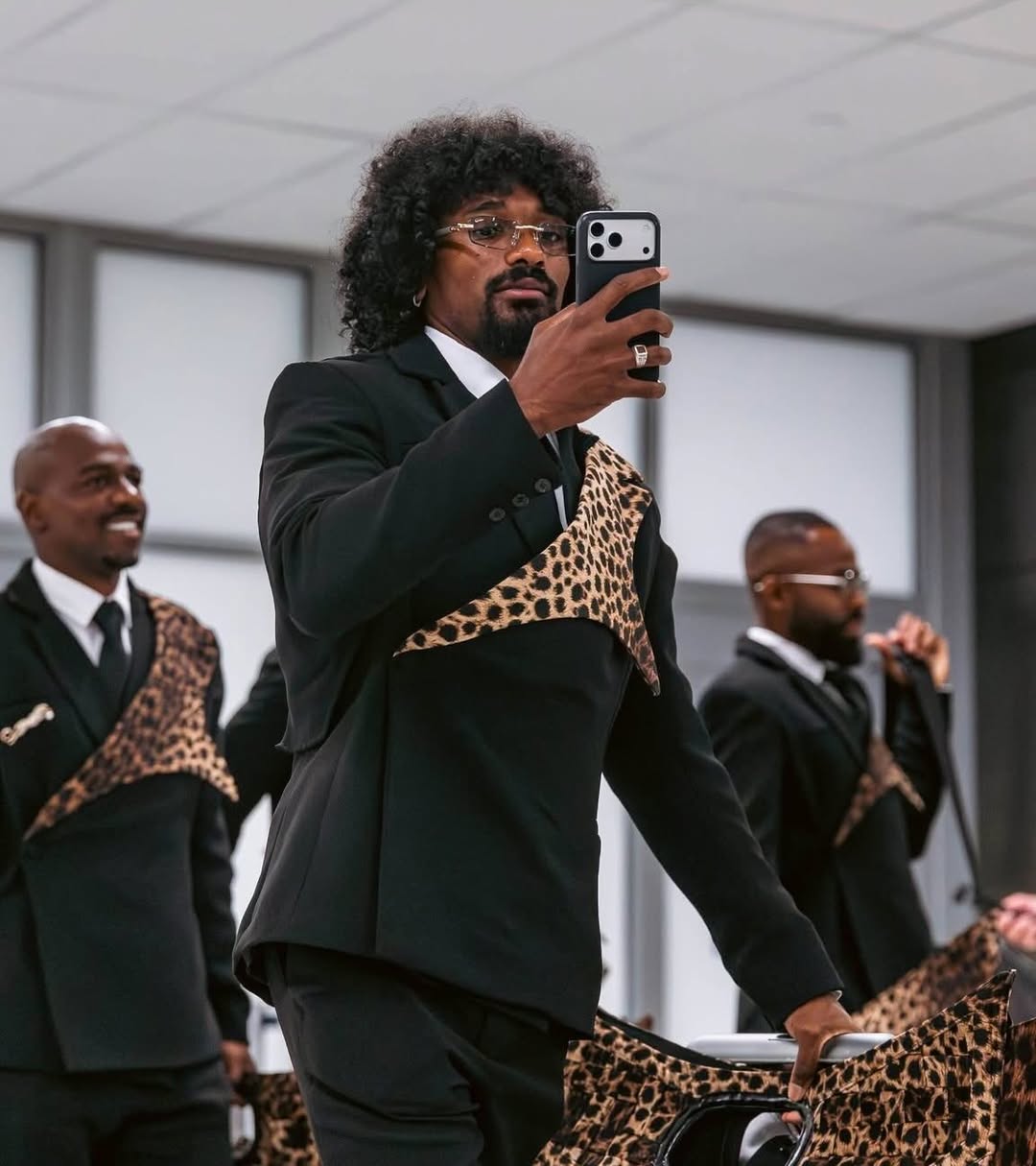DR Congo player with an afro hairstyle recording a video on his phone while walking in his official World Cup suit jacket.
