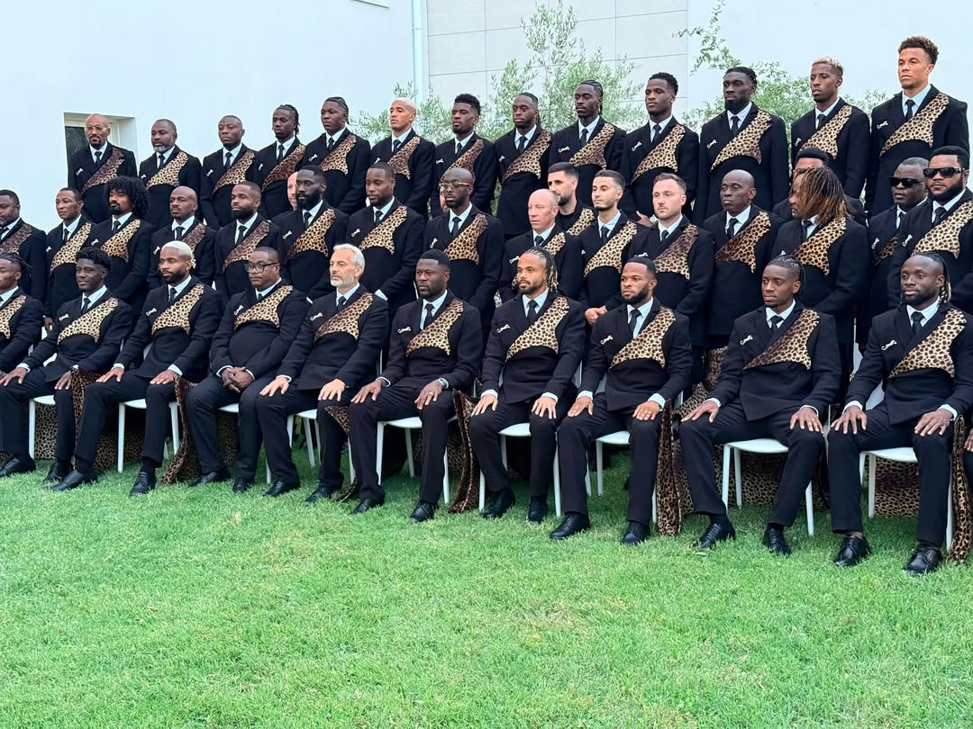 The DR Congo national football team squad seated in rows on grass wearing matching leopard-print panelled suits.