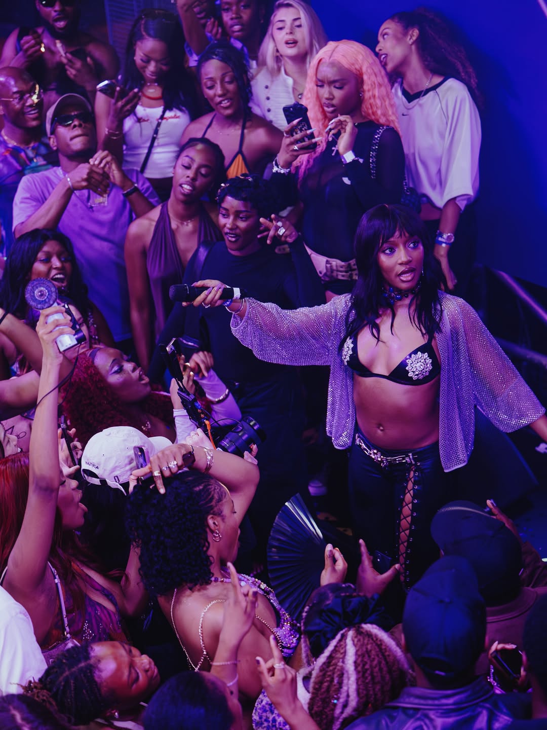 Ayra Starr holding a microphone and singing in a metallic mesh jacket and embellished bikini top surrounded by a crowd at her listening party.