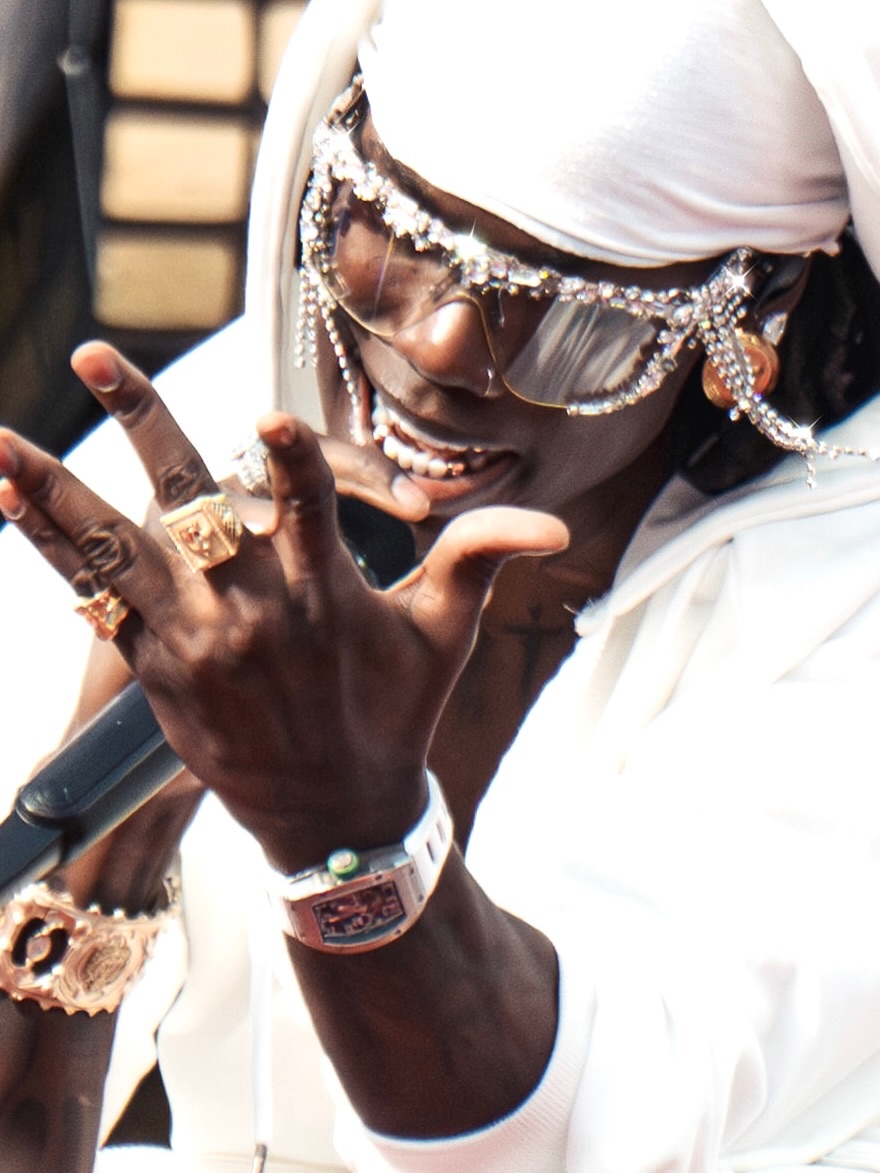 Rema gestures with his hands, displaying his gold rings, luxury watch, and futuristic sunglasses detailed with hanging crystal fringes.