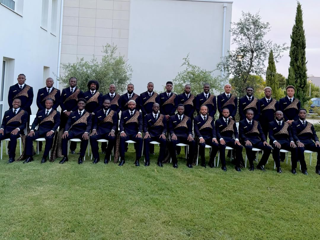 Full squad of DR Congo football team seated outdoors in uniform black suits featuring gold leopard pins and animal-print sashes.