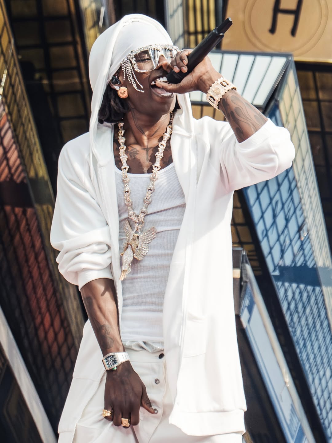 Rema holds a microphone to his mouth while wearing a white hooded jacket, a matched white wrap, and his angel chain pendant.