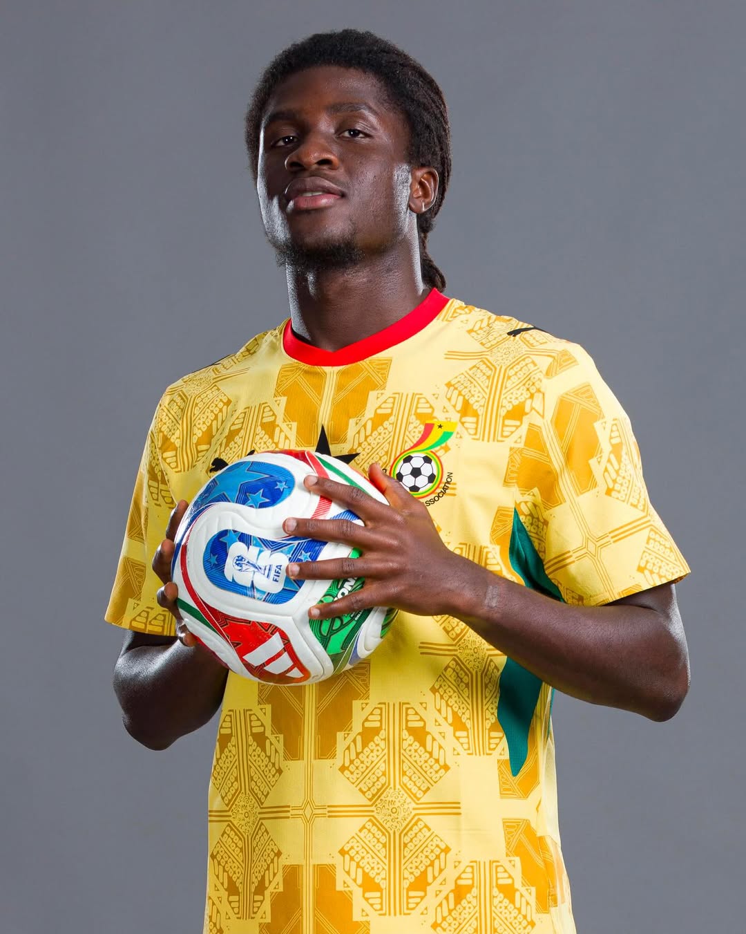 Studio portrait of Ghana national team football player Caleb Marfo Yirenkyi holding a FIFA match ball while wearing the official yellow and green patterned kit.