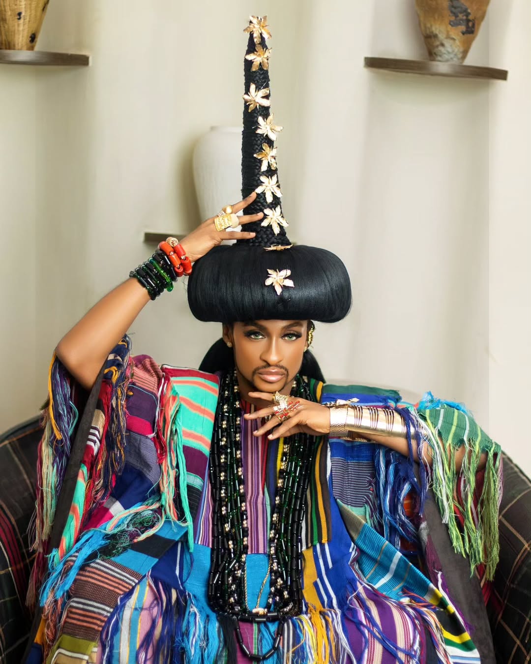Denrele Edun seated indoors wearing his multi-coloured woven Aso-Oke outfit, adjusting his tall black spire headpiece adorned with gold floral elements, displaying stacked bracelets and dark heavy beads.