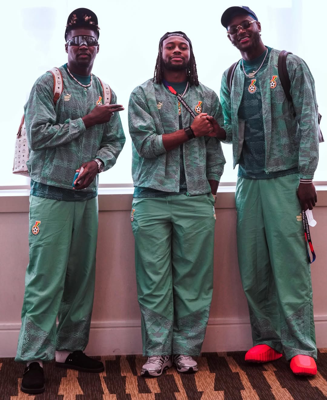 Jerome Opoku, Antoine Semenyo, and Joseph Anang, standing together indoors during their arrival in Canada, dressed in matching green patterned Puma travel tracksuits and sports gear.