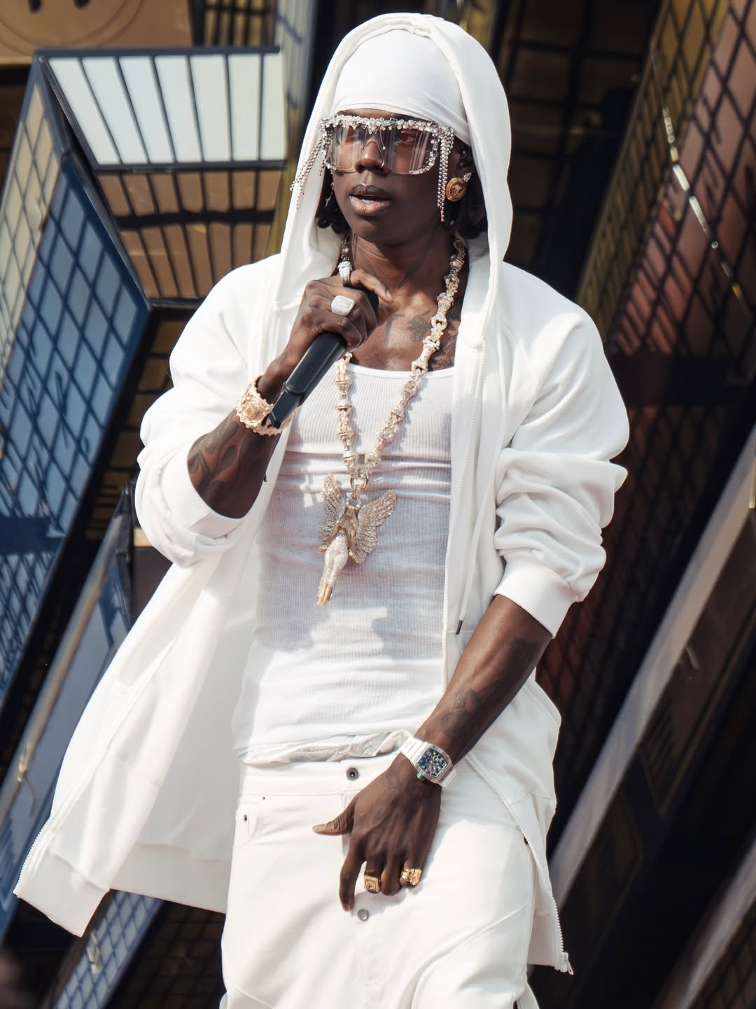 Rema stands on the outdoor stage holding a microphone, wearing his signature white hooded jacket, custom diamond eyewear, and rings.