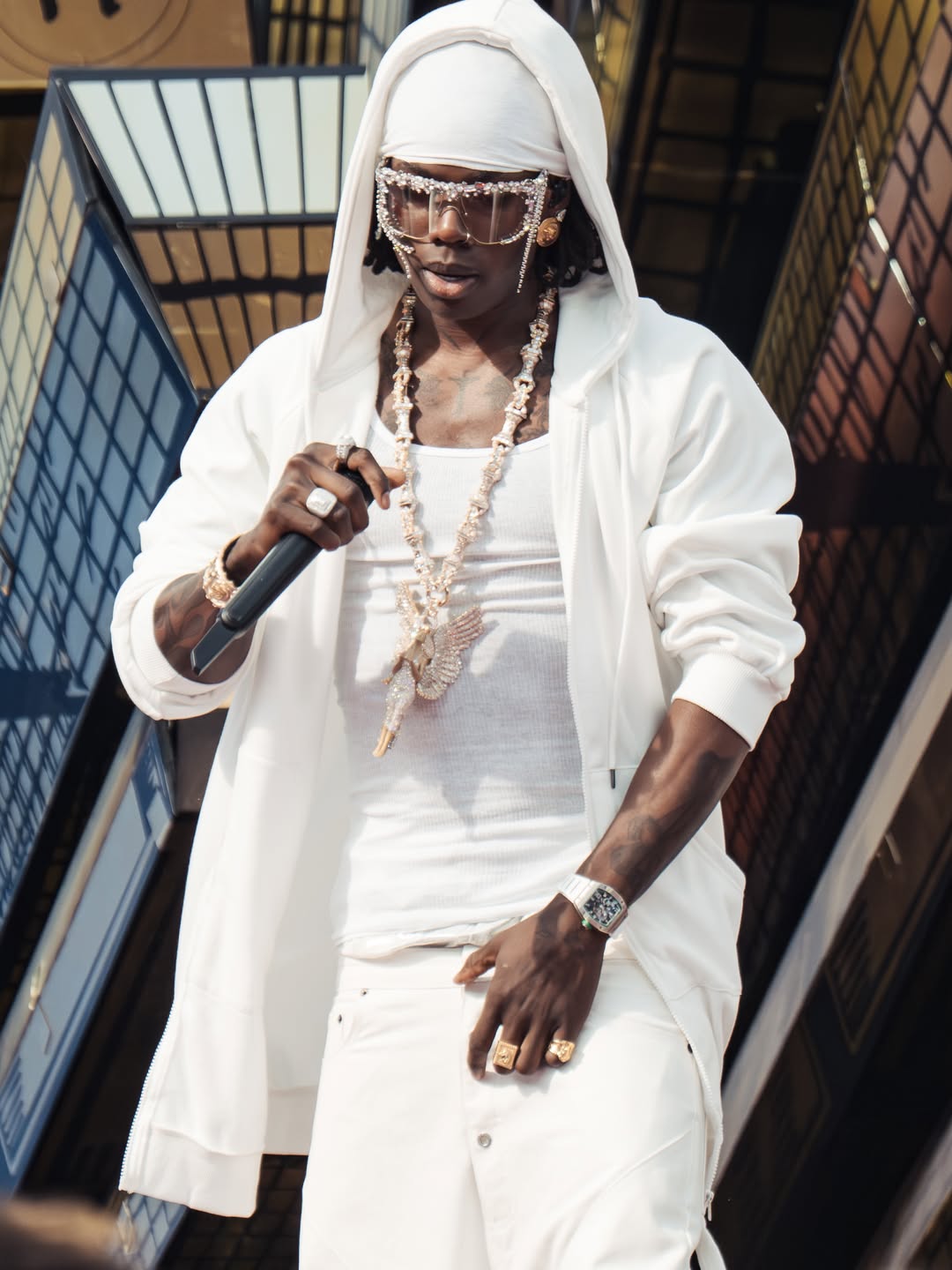 Rema sings into a microphone during his 2026 World Cup performance wearing a white hooded jacket, fringed diamond sunglasses, and a luxury watch