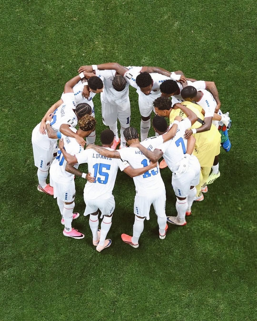 Overhead view of the Cabo Verde starting eleven in white jerseys gathered in a tight team huddle on the green grass.