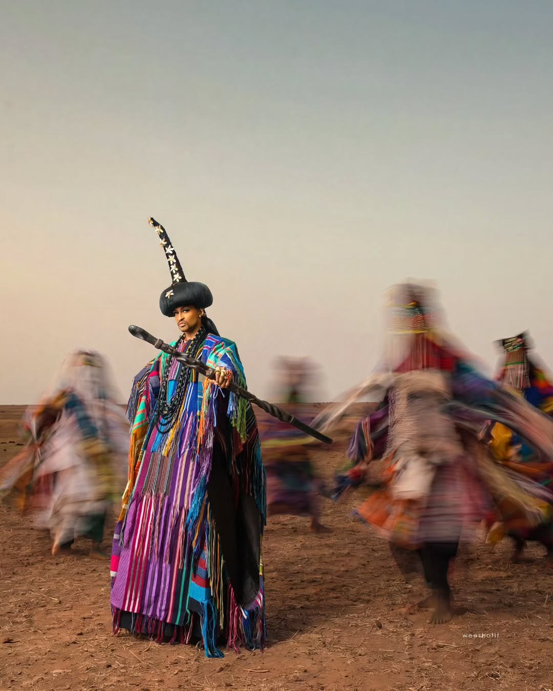 Denrele Edun posing in his birthday Aso-Oke garment made of vibrant purple, turquoise, and green panels, holding a carved wooden scepter across his chest against an open sky.