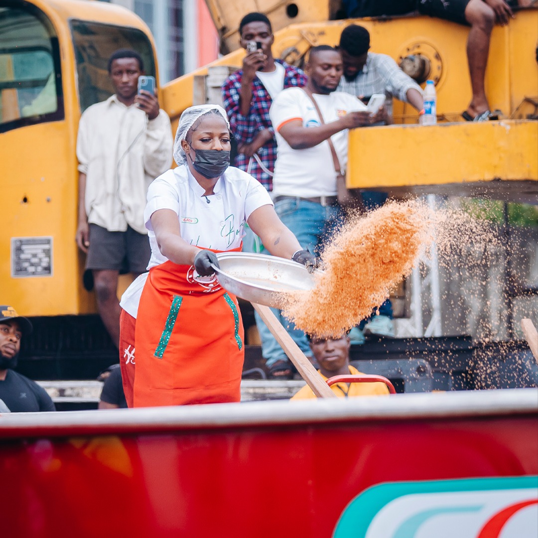Hilda Baci wearing a face mask and an orange apron while tipping ingredients from a large metal pan into a massive cooking pot during her second successful record run.