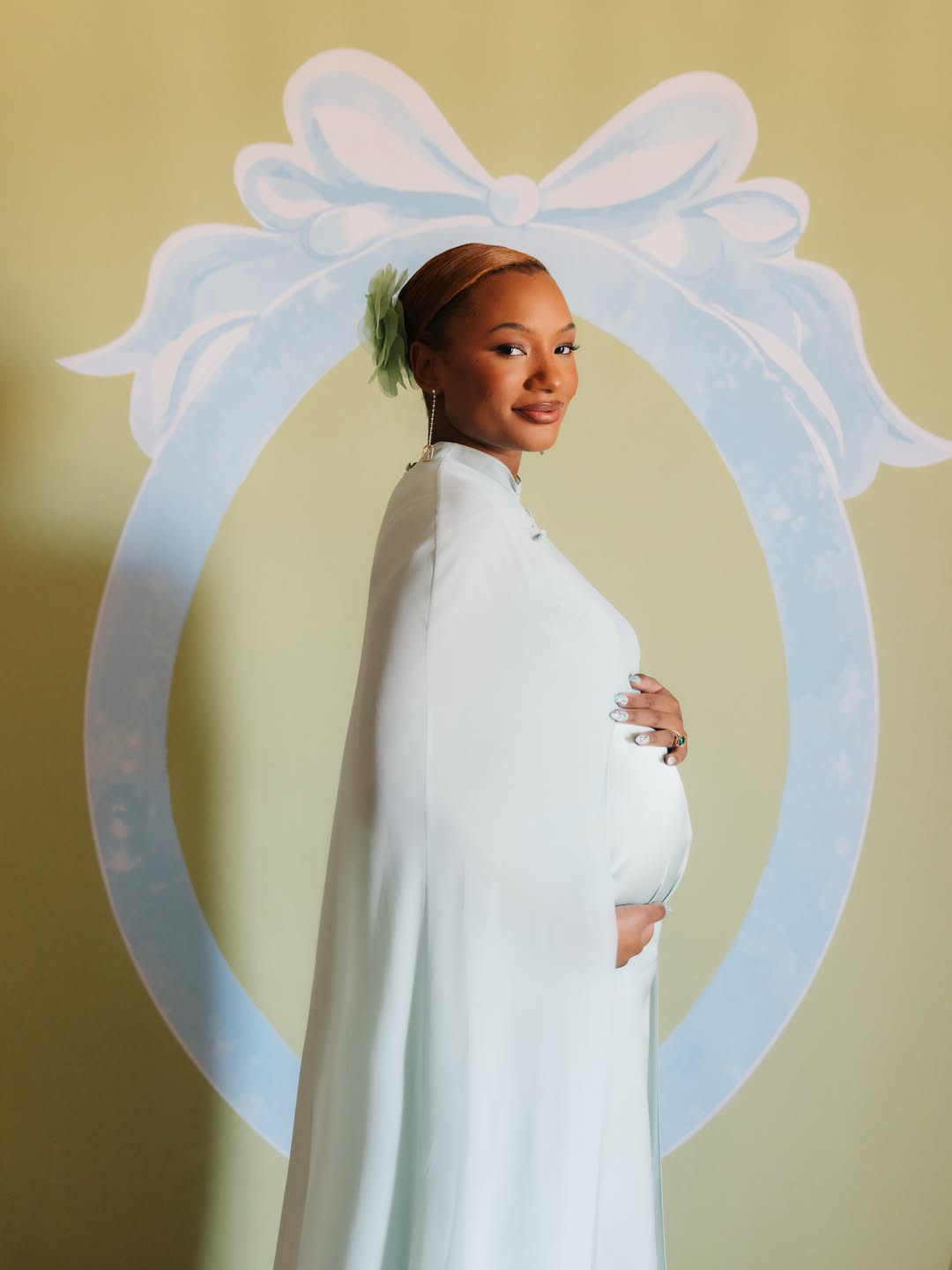 : Temi Ajibade cradling her pregnancy baby bump while wearing a mint-tinted ice blue silk maternity gown with a matching cape at her swan-themed baby shower.