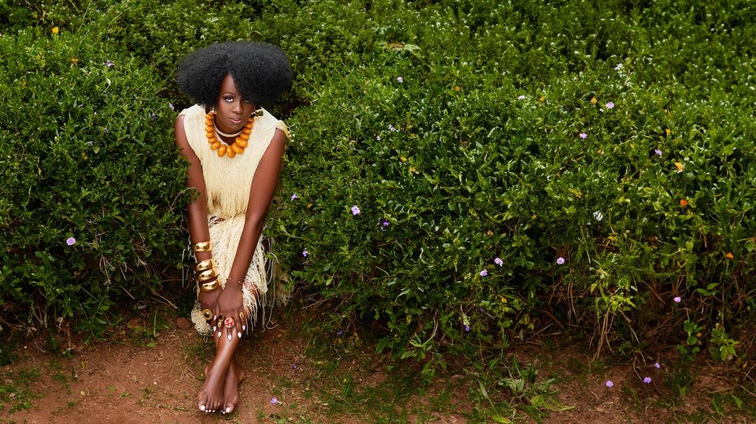 Singer Somi poses in a textured cream fringe dress and large orange bead necklace surrounded by green bushes.