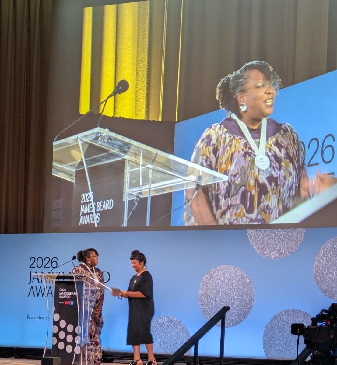 Ozoz Sokoh accepting her award at the podium during the 2026 James Beard Media Awards ceremony at the Art Institute of Chicago, with her image projected on the large screen above.