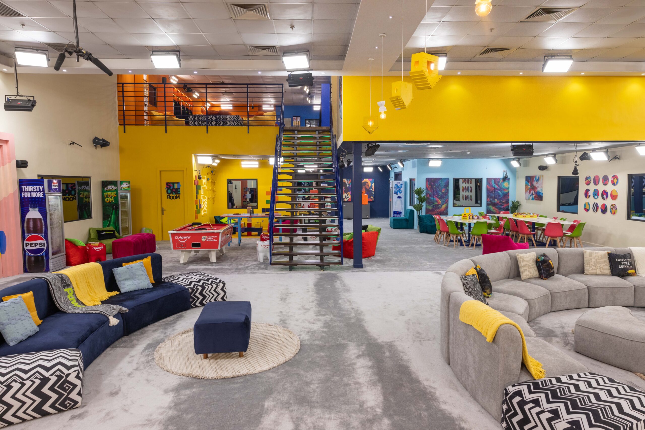 Open-plan main living room in the Big Brother Naija Season 11 house with curved grey sofas, blue seating, staircase, and gaming lounge