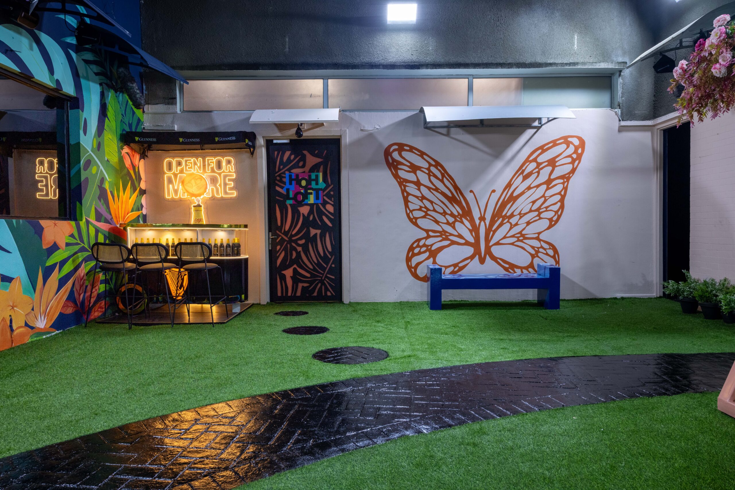 Outdoor garden bar in the Big Brother Naija Season 11 house with an orange butterfly mural and Open For More neon sign
