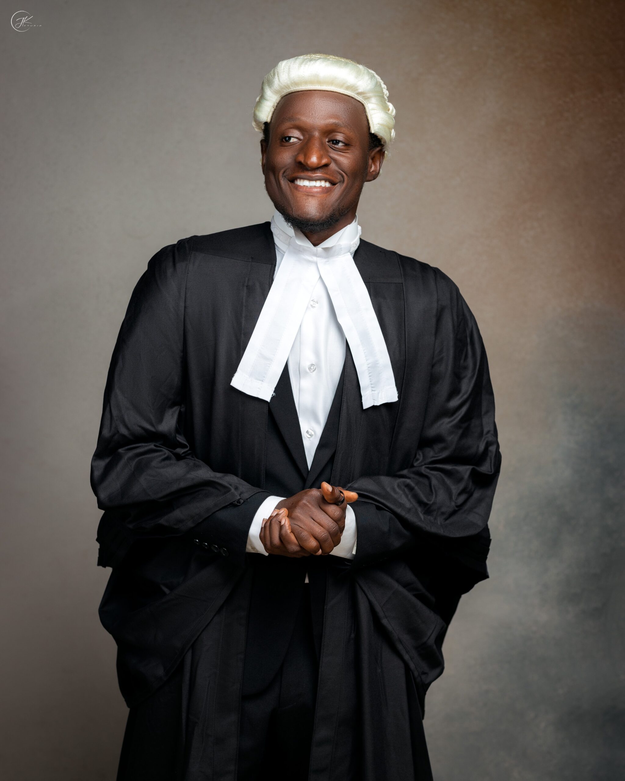 Nehemiah Shanum Danjuma in a black suit, white legal neckbands, and a traditional blonde-white barrister wig next to a hanging ceremonial gown.