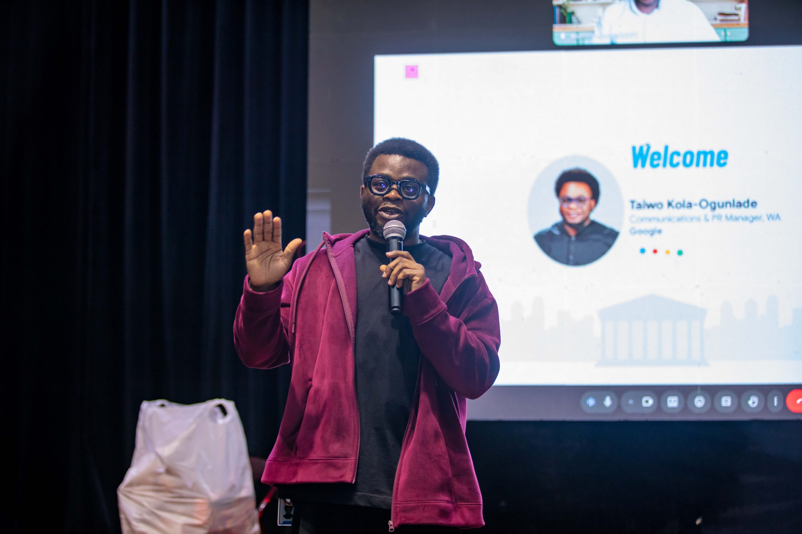 Taiwo Kola-Ogunlade wearing glasses and a maroon hoodie speaking with one hand raised in front of a digital screen displaying his introduction slide.