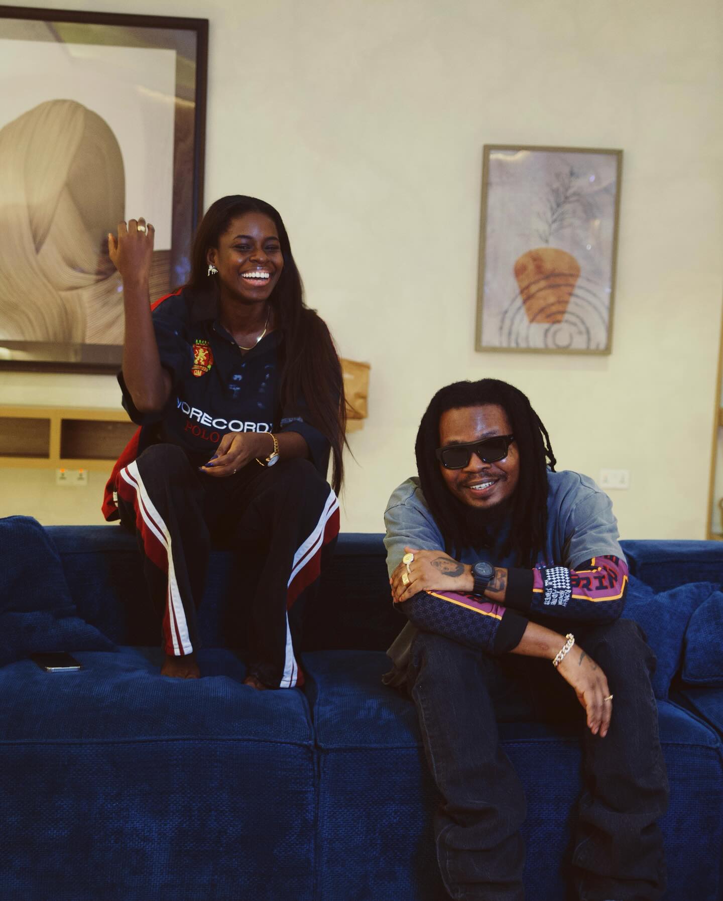 Nigerian YouTuber Korty EO laughing next to music artist Olamide on a blue couch during an interview in Lagos.