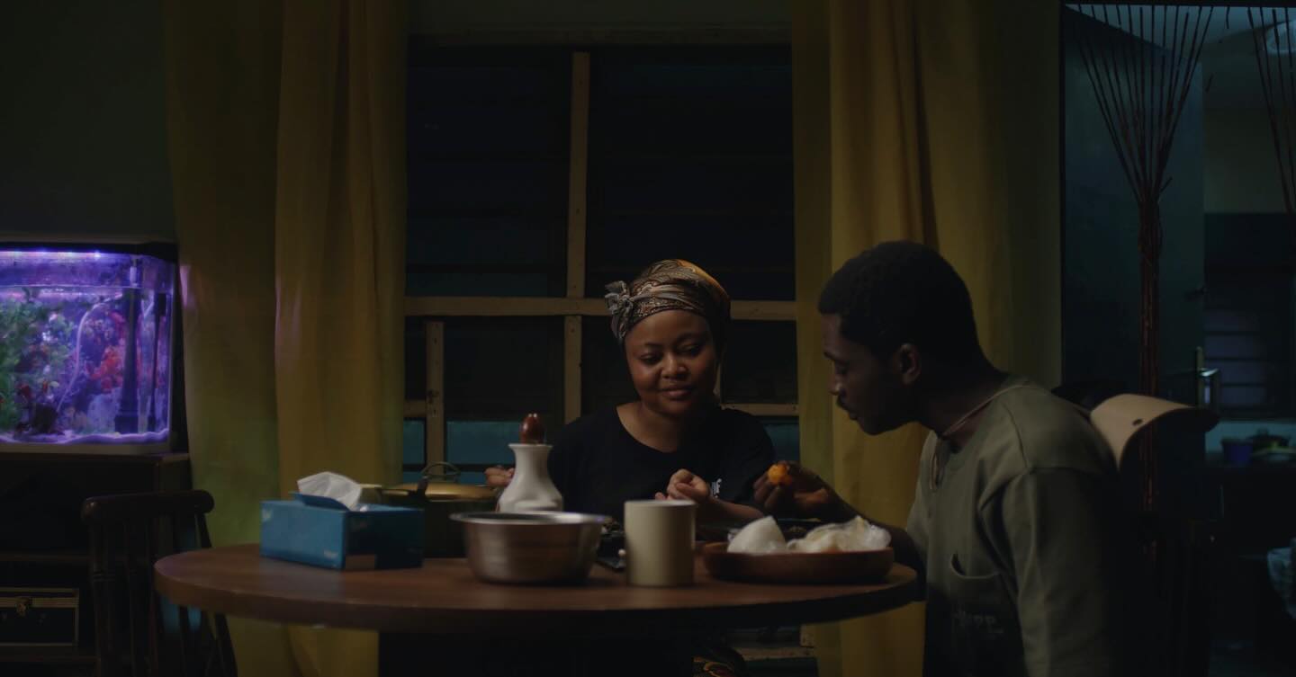 Teniola Aladese and a man having dinner at a wooden table next to a fish tank in the short film We Must Begin Again Together.