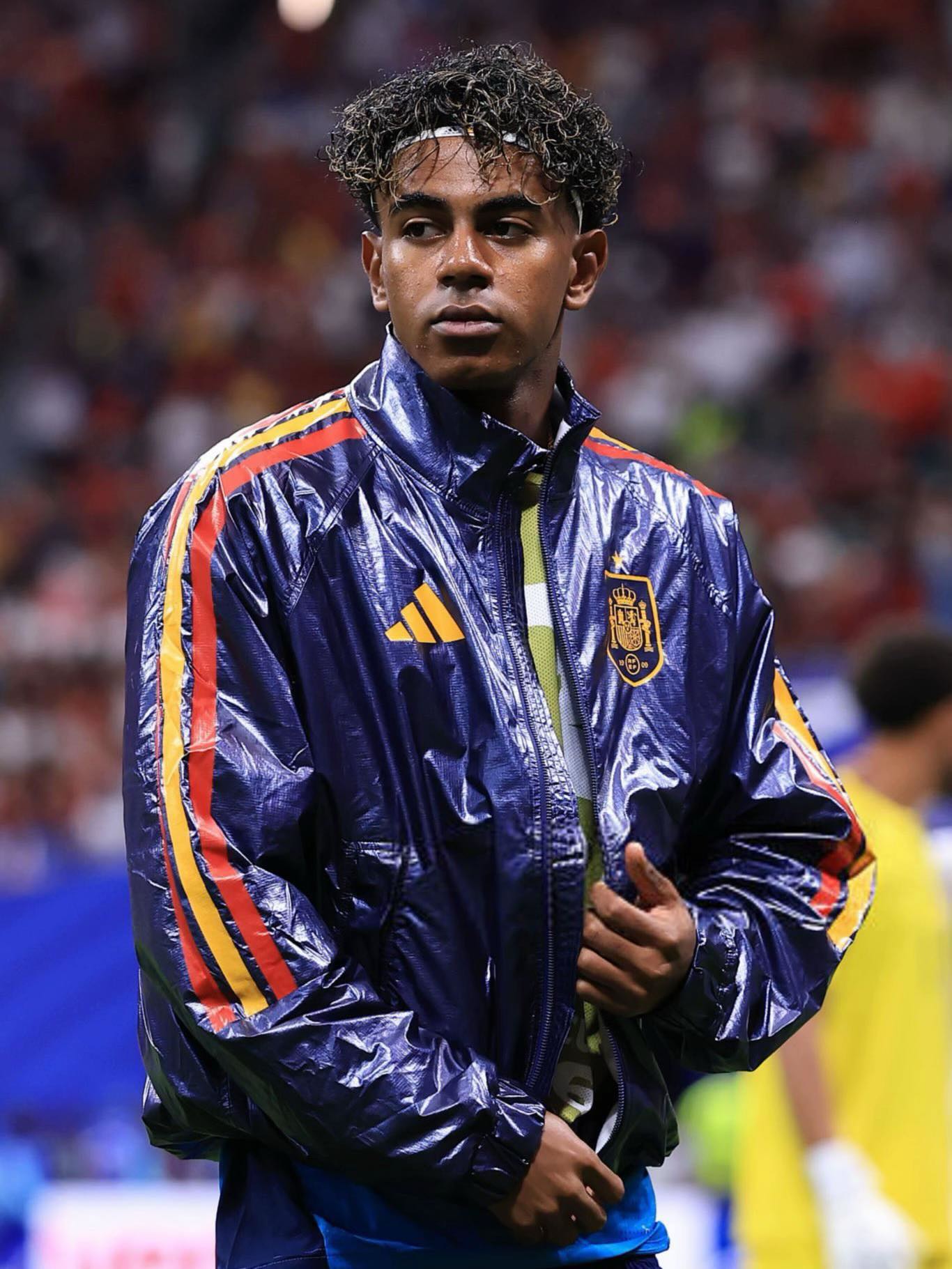 Lamine Yamal of Moroccan and Equatorial Guinean heritage looking sideways while wearing a glossy blue Spain national team anthem jacket on the pitch.